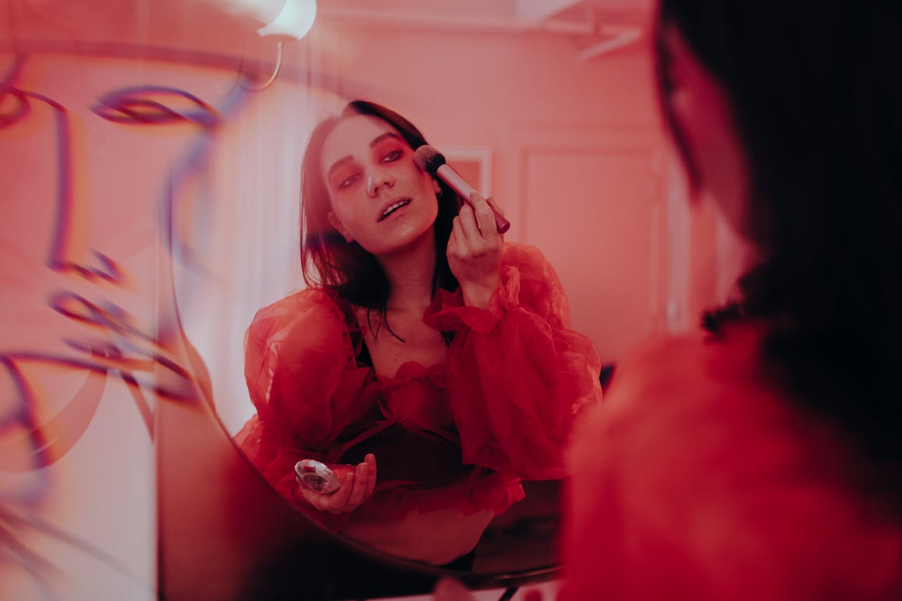 A woman applies makeup in front of a mirror reflecting an artistic red ambiance, creating a stylish, creative scene.